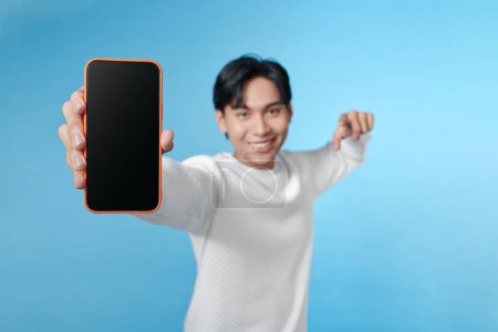 Téléchargez les photos : Joyeux homme pointent doigt tenir téléphone espace vide afficher écran isolé sur fond bleu - en image libre de droit