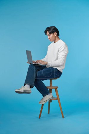 Téléchargez les photos : Beau homme émotionnel assis sur des tabourets et utilisant un ordinateur portable isolé sur fond bleu - en image libre de droit