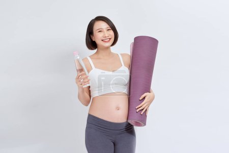 Téléchargez les photos : Happy fitness femme enceinte tenant tapis de yoga et bouteille d'eau isolé sur fond blanc - en image libre de droit