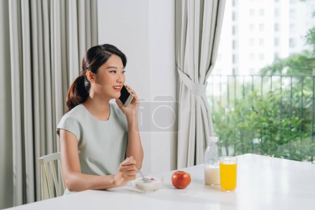 Téléchargez les photos : Jolie femme assise à la cuisine, mangeant des flocons d'avoine et appelant - en image libre de droit