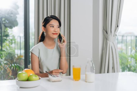 Téléchargez les photos : Femme utilisant un téléphone portable tout en prenant le petit déjeuner à la maison
. - en image libre de droit