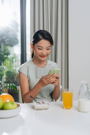 Foto de Mujer joven desayunando saludable mientras usa el teléfono inteligente en casa - Imagen libre de derechos