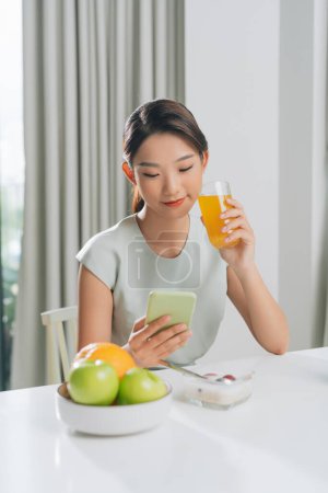 Téléchargez les photos : Belle femme buvant du jus d'orange et lisant les nouvelles en ligne dans la cuisine - en image libre de droit