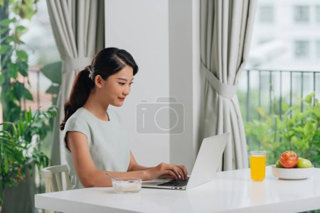 Téléchargez les photos : Belle jeune femme travaillant sur ordinateur portable tout en étant assis à la table du petit déjeuner le matin
. - en image libre de droit