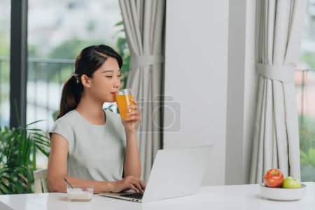 Téléchargez les photos : Femme asiatique positive avec du jus d'orange en utilisant un ordinateur portable dans la cuisine - en image libre de droit