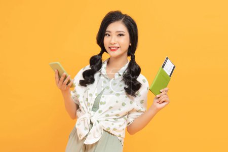 Foto de Niza sonriente mujer asiática con pasaporte con billete y el uso de teléfono celular sobre fondo amarillo - Imagen libre de derechos