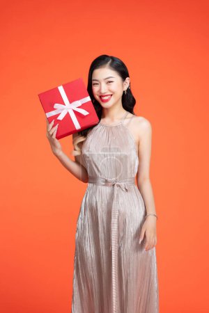 Téléchargez les photos : Jeune femme portant une robe brillante tenant une boîte-cadeau rouge devant une toile de fond rouge - en image libre de droit