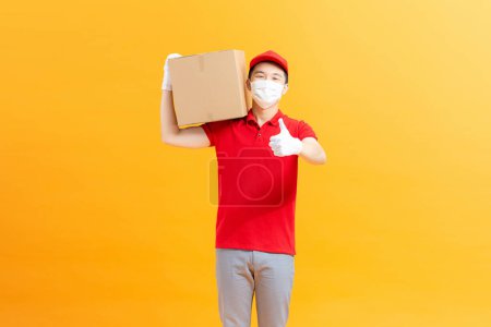Téléchargez les photos : Jeune homme de livraison asiatique portant un masque de protection et des gants donnant du carton de colis au client - en image libre de droit