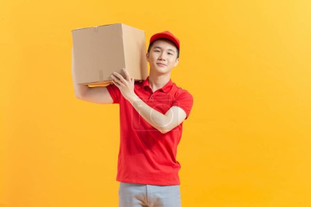 Téléchargez les photos : Amical asiatique livraison gars portant chemise rouge et casquette avec boîte colis sur fond jaune - en image libre de droit