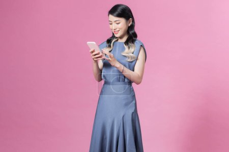 Téléchargez les photos : Jeune femme portant une robe en soie bleue à l'aide d'un smartphone sur fond rose - en image libre de droit