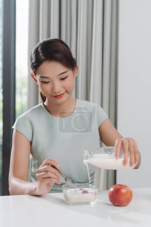 Téléchargez les photos : Femme heureuse versant du lait frais du verre dans un bol avec des céréales, avoir un délicieux repas - en image libre de droit