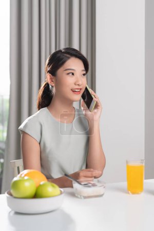 Foto de Hermosa chica asiática sonriente hablando por teléfono sentado en la mesa en el comedor en la mañana - Imagen libre de derechos
