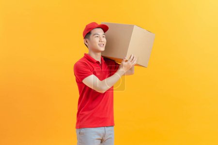 Photo for Asian delivery man in red uniform with parcel cardboard box isolated on yellow background - Royalty Free Image