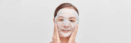 Téléchargez les photos : Femme appliquant un masque de feuille sur son visage, sur une bannière blanche - en image libre de droit
