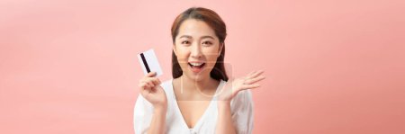 Téléchargez les photos : Orientation panoramique d'une femme asiatique gaie tenant sa carte de crédit isolée sur rose - en image libre de droit