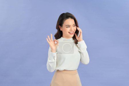 Photo for Cheerful Asian woman holding smartphone and shows ok sign on light violet background. - Royalty Free Image
