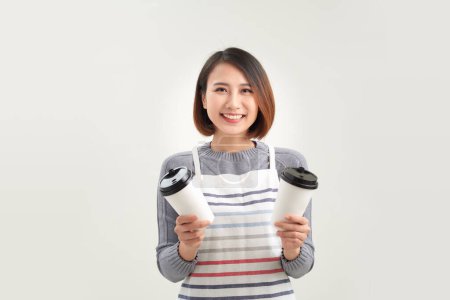 Foto de Amistoso asiático joven mujer en delantal celebración takeaway papel café tazas - Imagen libre de derechos