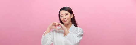 Téléchargez les photos : Heureux sourire jeune femme faisant geste de coeur de main sur fond rose - en image libre de droit