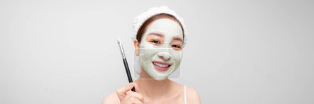 Téléchargez les photos : Joyeux jeune femme appliquant un masque blanc sur son visage avec un pinceau. - en image libre de droit