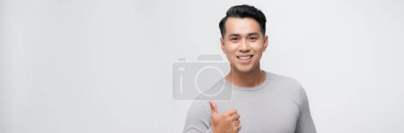 Foto de Joven guapo asiático hombre sobre blanco banner fondo haciendo feliz pulgar hacia arriba gesto con mano. - Imagen libre de derechos