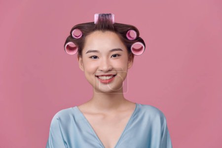 Foto de La ama de casa aplica rulos para el cabello, vestida con bata casual, posa sobre un fondo rosado - Imagen libre de derechos