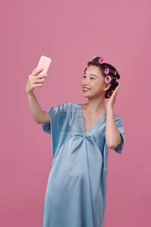 Téléchargez les photos : Jeune femme en robe de soie avec des bigoudis prenant selfie sur fond rose - en image libre de droit