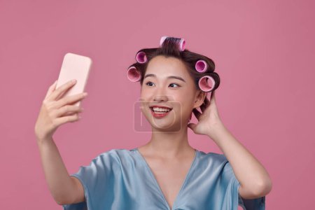 Téléchargez les photos : Joyeux jeune femme en robe décontractée habillée avec des bigoudis de cheveux prenant selfie sur fond rose - en image libre de droit