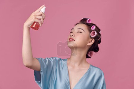 Téléchargez les photos : Belle femme asiatique appliquant de l'eau de pulvérisation sur le visage sur fond rose avec des bigoudis dans ses cheveux - en image libre de droit