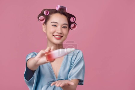 Téléchargez les photos : Jeune femme souriante dans les bigoudis tenant bouteille cosmétique pulvérisation à la main et près du visage - en image libre de droit