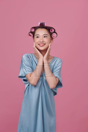Photo for Housewife applies hair curlers, dressed in casual robe, poses against rosy background - Royalty Free Image