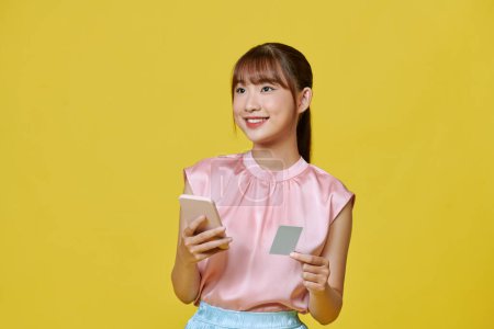 Photo for Happy young asian woman using mobile phone and credit card isolated over yellow background. - Royalty Free Image
