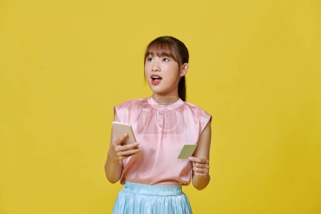 Téléchargez les photos : Heureux jeune asiatique femme en utilisant téléphone mobile et carte de crédit isolé sur fond jaune. - en image libre de droit