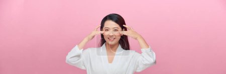 Foto de Hermosa modelo femenina asiática sonriente con maquillaje natural tocando la piel hidratada brillante sobre fondo rosa. - Imagen libre de derechos