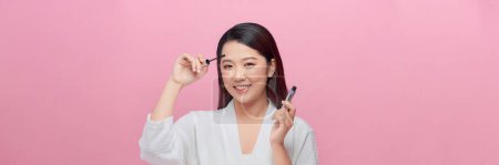 Foto de Mujer joven y feliz aplicando rímel con cepillo en las pestañas mientras hace maquillaje sobre fondo rosa - Imagen libre de derechos