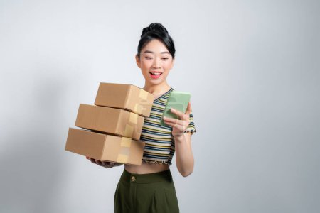 Photo for Young happy asian woman wear summer casual clothes hold stack cardboard blank boxes use mobile  phone - Royalty Free Image