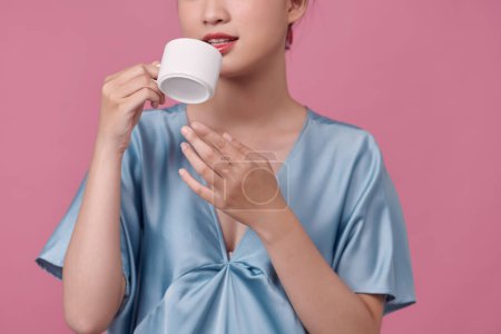 Téléchargez les photos : Joyeux jeune femme en robe de chambre et bigoudis de cheveux boire du café sur fond rose - en image libre de droit