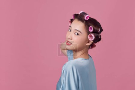Téléchargez les photos : Jeune femme incertaine et souriante sans maquillage et avec des bigoudis sur rose - en image libre de droit