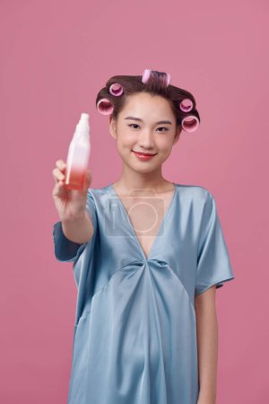 Téléchargez les photos : Belle jeune femme tenant une bouteille dans sa main avec des bigoudis sur ses cheveux - en image libre de droit
