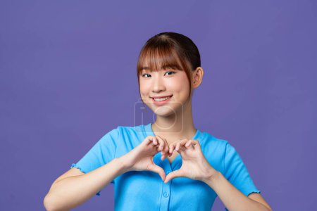 Photo for Young asian woman shows her fingers heart-shaped sign and presses to chest on purple background. - Royalty Free Image