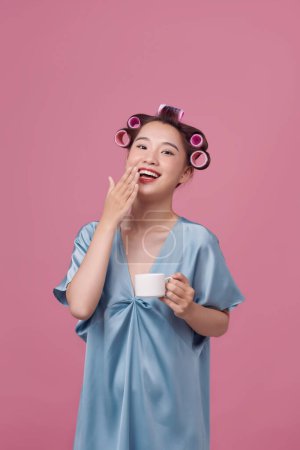 Foto de Hermosa mujer joven en vestido azul y rizadores de pelo bebiendo café sobre fondo rosa - Imagen libre de derechos