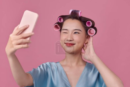 Foto de Mujer alegre aplica rulos de pelo posando en cámara smartphone vestido sobre fondo rosa. - Imagen libre de derechos