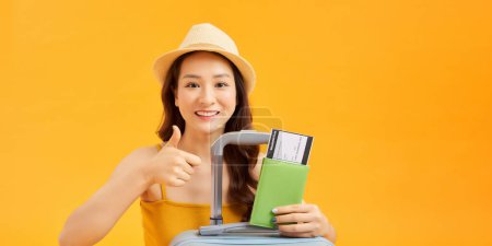 Photo for Dreamy asian tourist, smiling and thinking, holding ticket and passport with suitcase for trip - Royalty Free Image