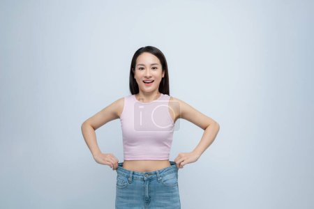 Foto de Exitosa pérdida de peso, mujer asiática con jeans demasiado grandes después de una dieta efectiva, fondo blanco - Imagen libre de derechos