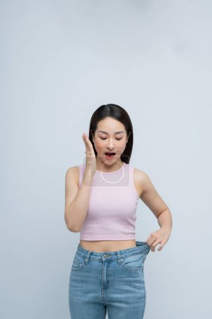 Foto de Exitosa pérdida de peso, mujer asiática con jeans demasiado grandes después de una dieta efectiva, fondo blanco - Imagen libre de derechos