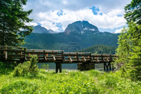 Photo for Wooden footbridge over stream against background of Meded Peak and Glacial Black Lake. Durmitor National Park. Walking path near lake is a popular destination for recreation and hiking. Montenegro. - Royalty Free Image