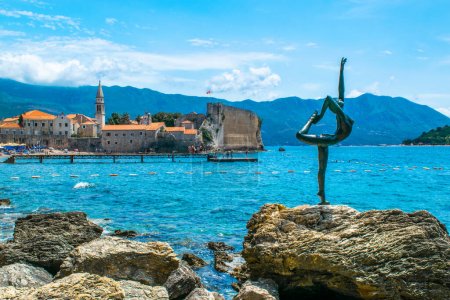 Foto de Budva, Montenegro - 4 de julio de 2021: Escultura de bailarina (Estatua de gimnasta o bailarina) sobre el fondo de la antigua fortaleza de las murallas de la ciudad. El casco antiguo de Budva. Hermoso azul verano soleado paisaje marino. - Imagen libre de derechos
