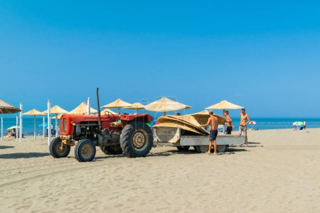 Photo for Ulcinj, Montenegro - 09 July 2021: Workers equip the sand Long Beach (Velika plaza) with umbrellas brought on a tractor. Sunny blue seaside landscape. Adriatic Sea. - Royalty Free Image