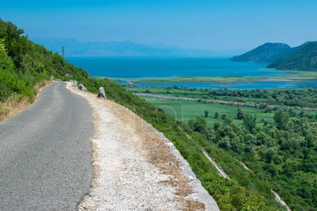 Schmale Straße in der Nähe des Skadar Sees mit grünem und blauem Wasser. Schöne Sommerlandschaft mit Bergen und Hügeln. Klarer Himmel. Montenegro. Reisekonzept.