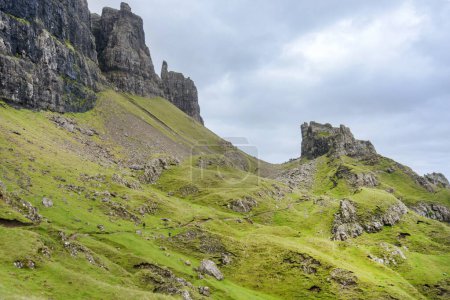 Foto de Hermoso y dramático paisaje montañoso escocés, picos de montaña puntiagudos y dentados y rostros escarpados de acantilados, a lo largo de la caminata de las colinas Quiraing, hierba verde y arbustos cubiertos a mediados del verano, al noreste de Skye. - Imagen libre de derechos