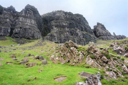 Photo for Sheer vertical sides of jagged dark rock faces,lining the Quraing escarpment,northernmost summit of the Trotternish,ancient landslide,mountain trail beneath covered with summer grass. - Royalty Free Image
